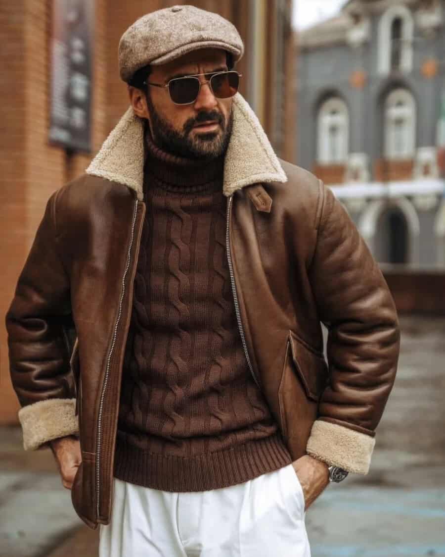 Man wearing white pants, brown cable knit turtleneck, brown shearling aviator jacket, square sunglasses and brown Cappellificio Biellese baker boy hat