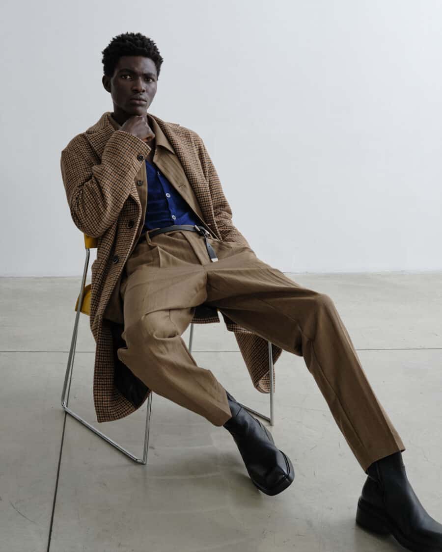Black man wearing Italian brand Barena brown pleated pants, blue shirt, brown tweed overcoat and black leather Chelsea boots