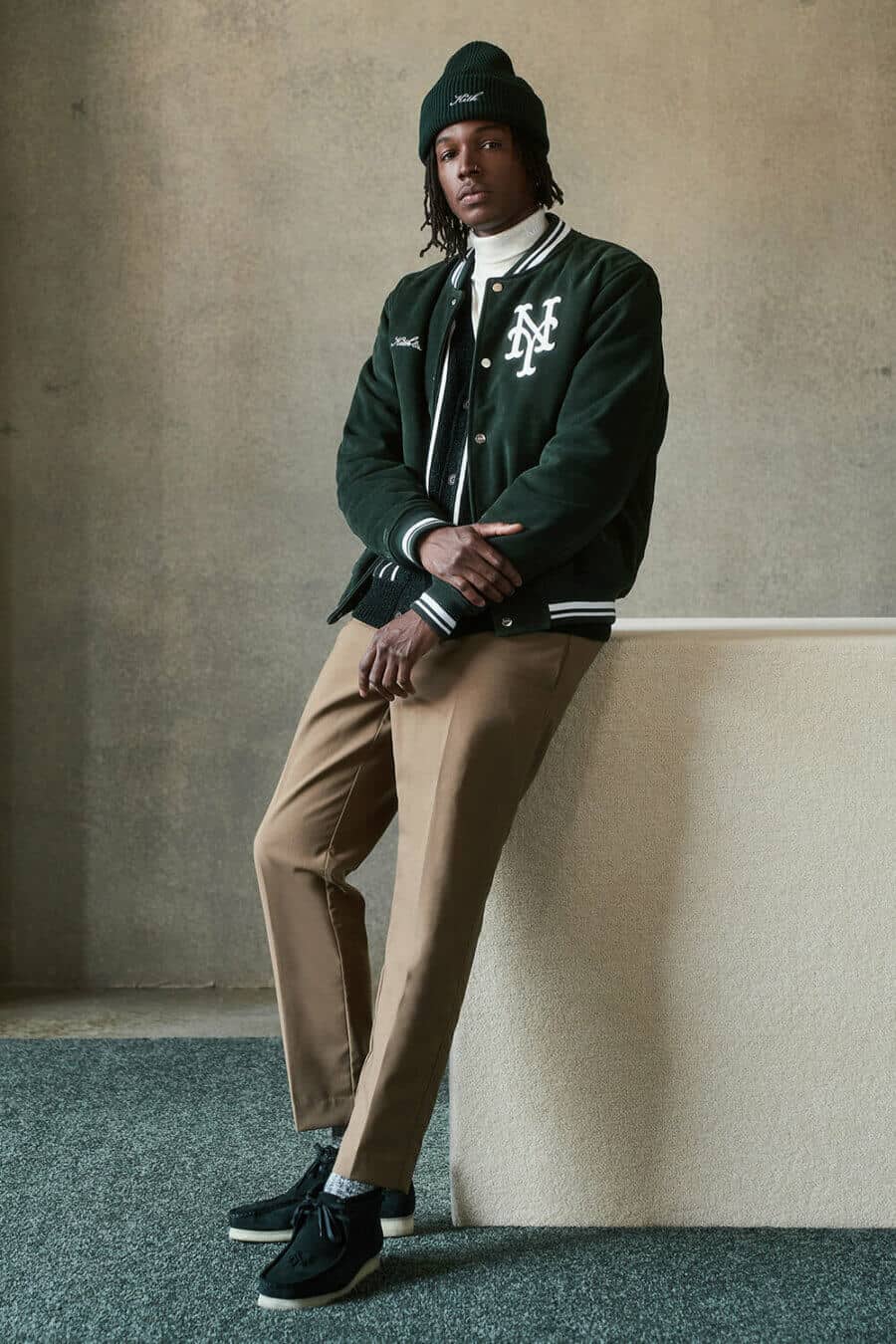Men's tailored khaki pants, white turtleneck, green varsity jacket, beanie and suede Wallabee boots outfit