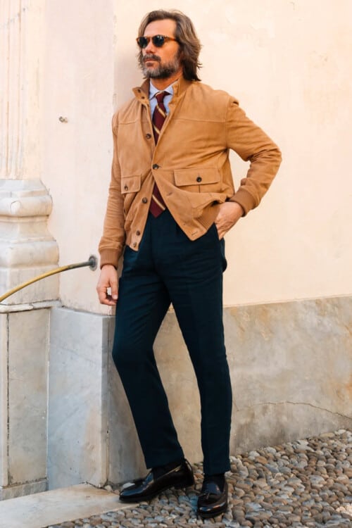 Italian man wearing navy pants, light blue shirt, club rep tie, tan suede bomber jacket, sunglasses and black leather tassel loafers