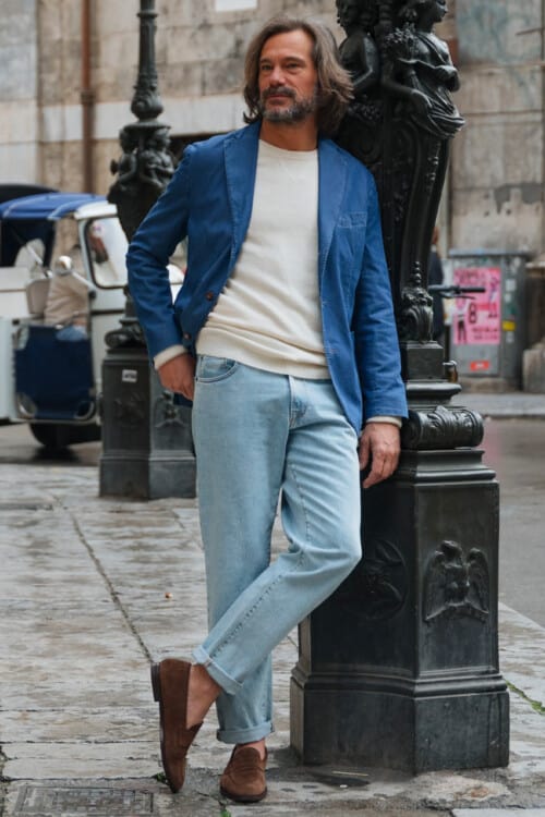 Italian man wearing light wash blue jeans, white long sleeve top, blue cotton twill blazer and brown suede penny loafers