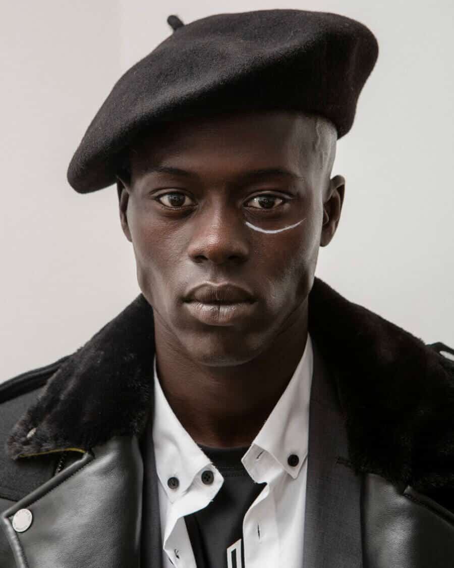 Black male model Alpha Dia wearing a white shirt, black beret hat and black leather fur collar biker jacket