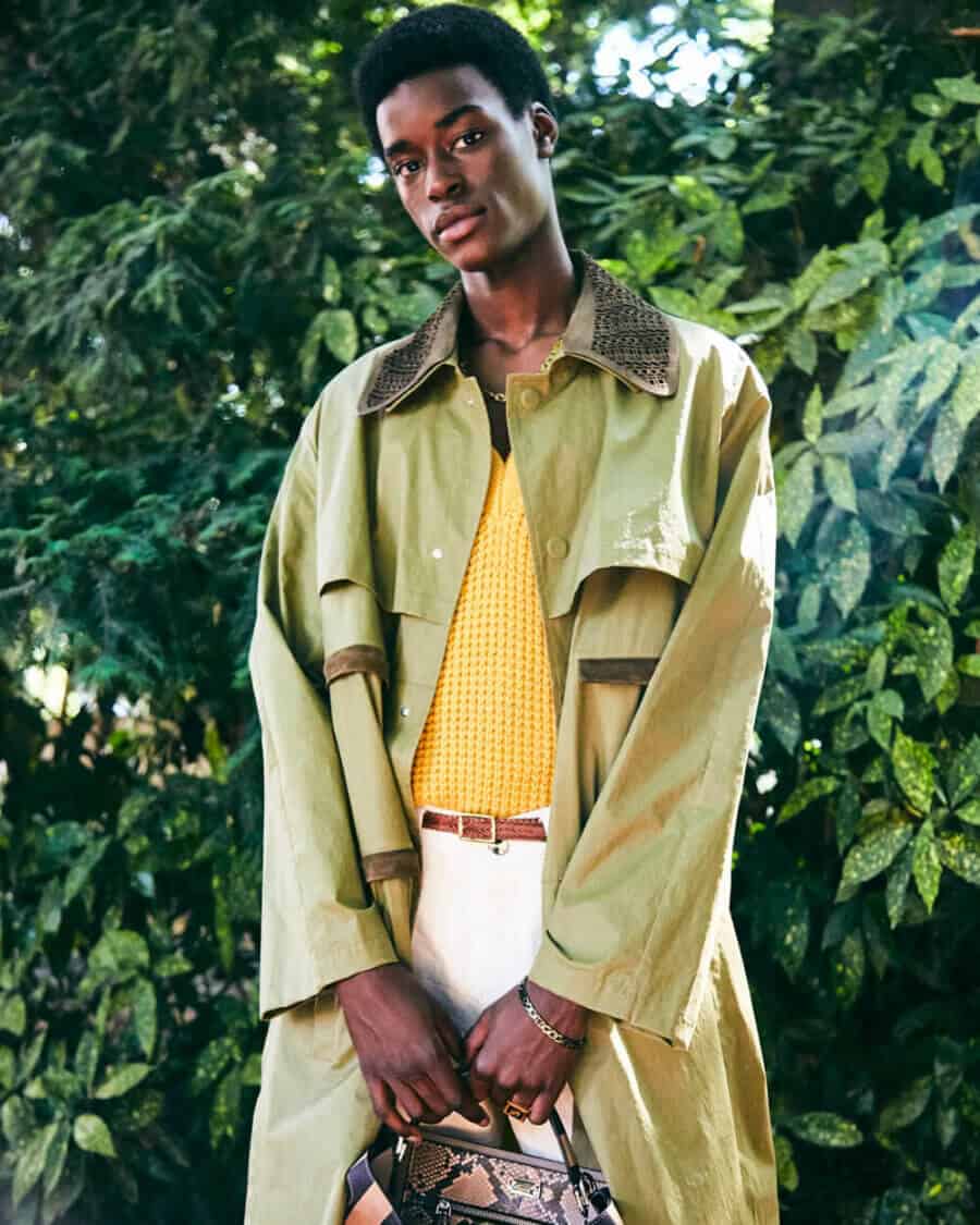 Black male model Babacar N’Doye wearing white pants, yellow crochet top and light green trench coat