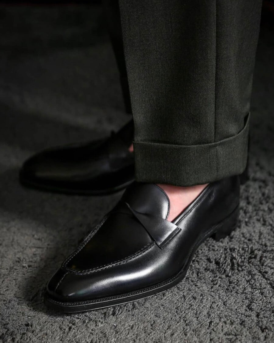 Man wearing Gaziano & Girling black leather pointed loafers on feet sockless with grey tailored turn-up pants