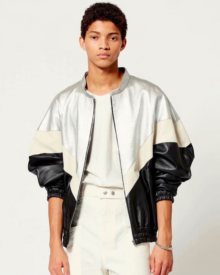 Black male model Jeranimo Van Russel wearing off-white pants, a tucked in off-white T-shirt and a silver, black and beige bomber jacket