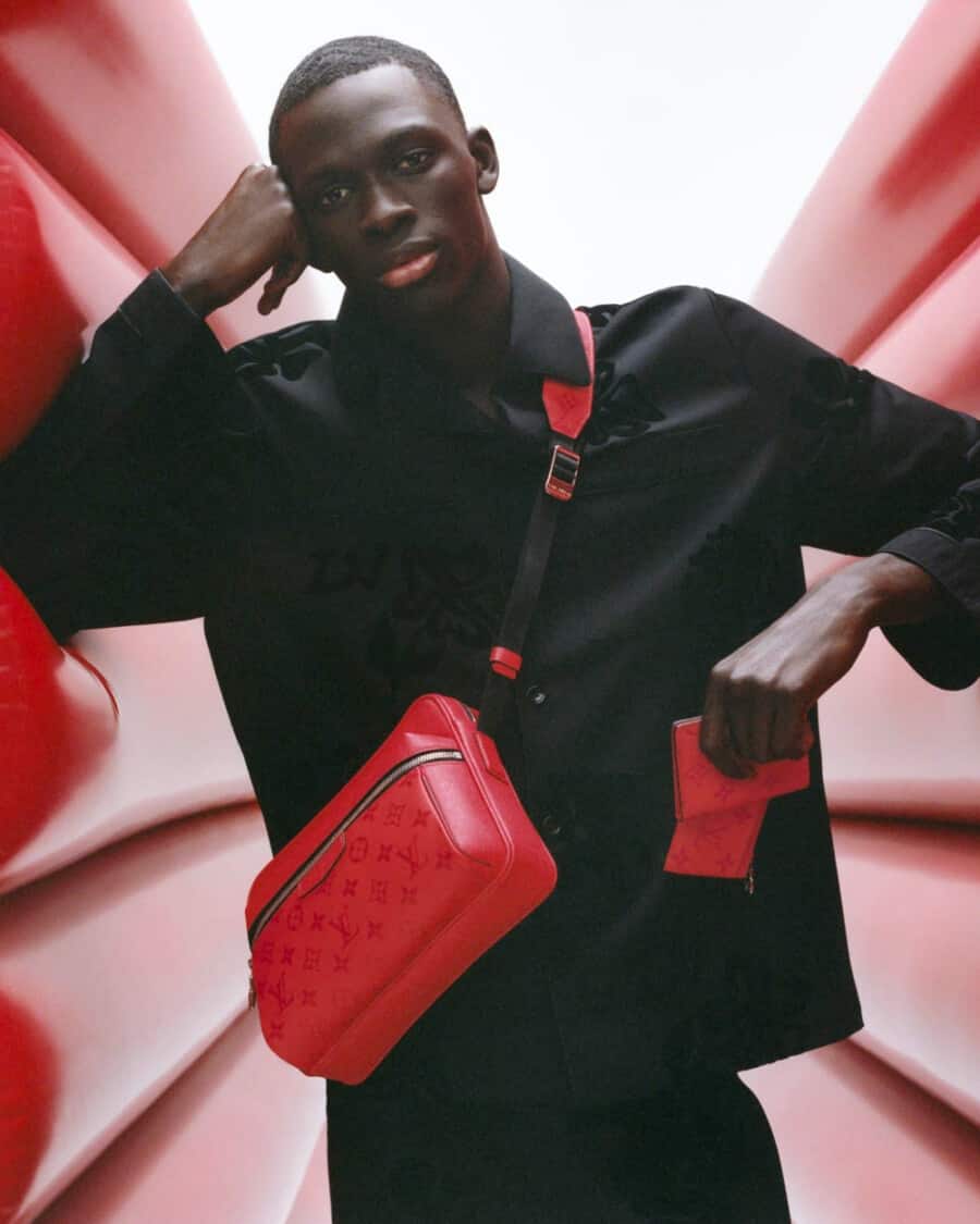 Black male model Khadim Sock wearing a black embroidered shirt and black pants with a bright red Louis Vuitton crossbody bag