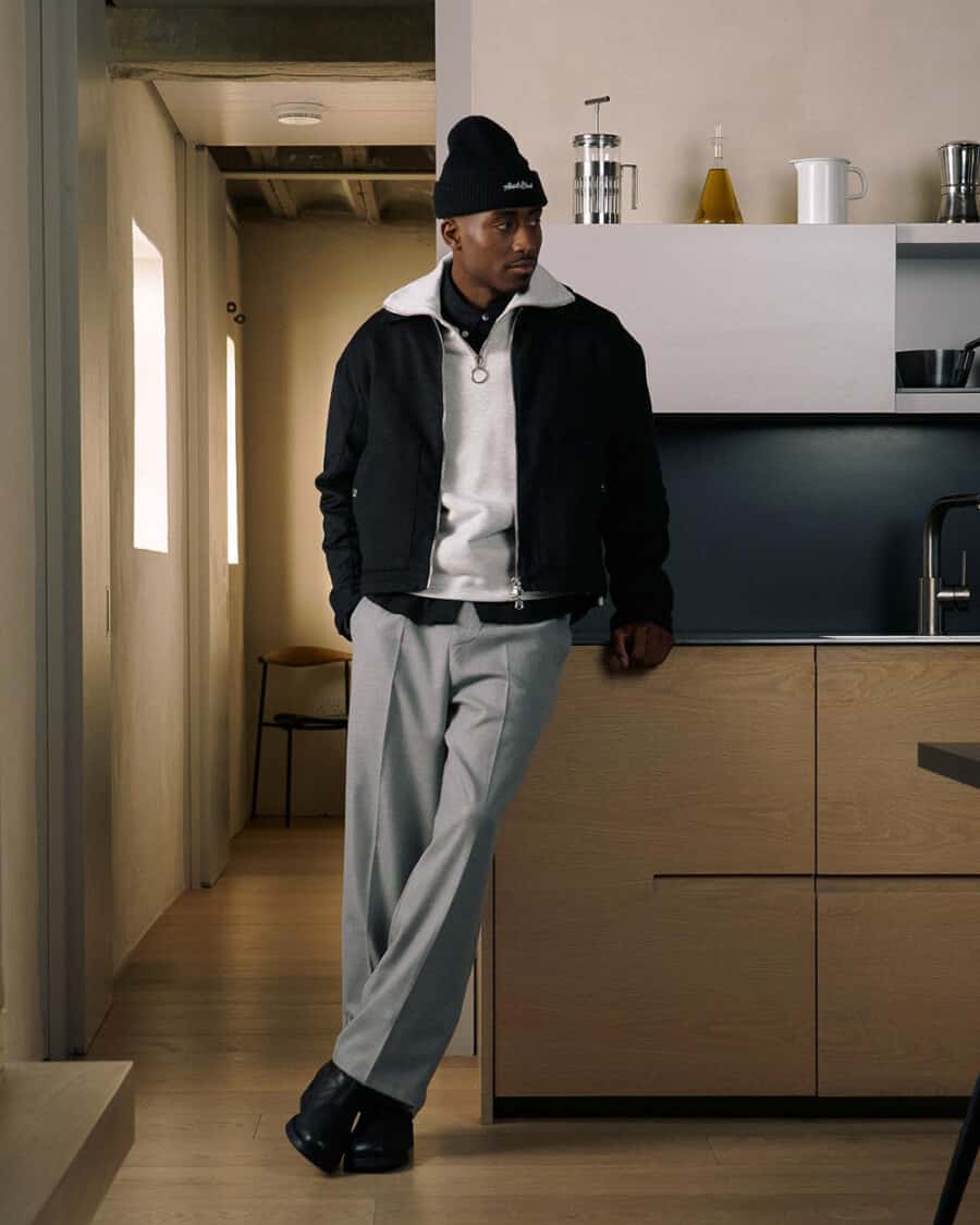 Man wearing about:blank streetwear grey pants, black shirt, grey quarter-zip sweatshirt, black bomber jacket, black beanie and black Chelsea boots