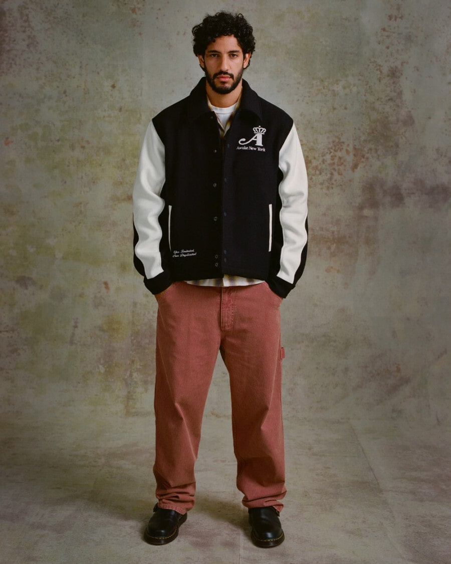 Man wearing AWAKE NY streetwear brown carpenter pants, brown-white checked shirt, black and white varsity jacket and black Dr Martens