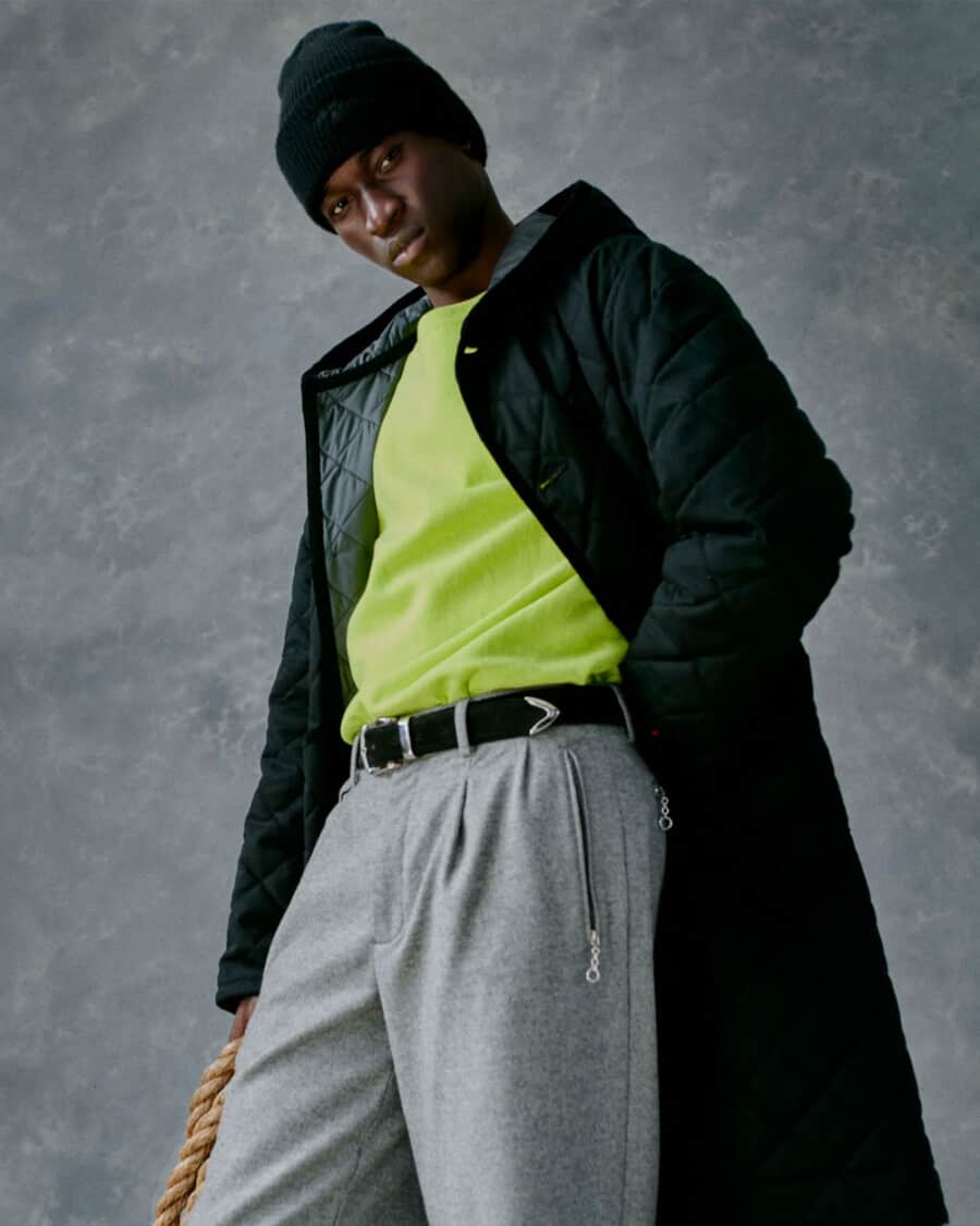 Man wearing Noah NY grey pleated pants, lime green T-shirt, quilted long length coat and green beanie streetwear