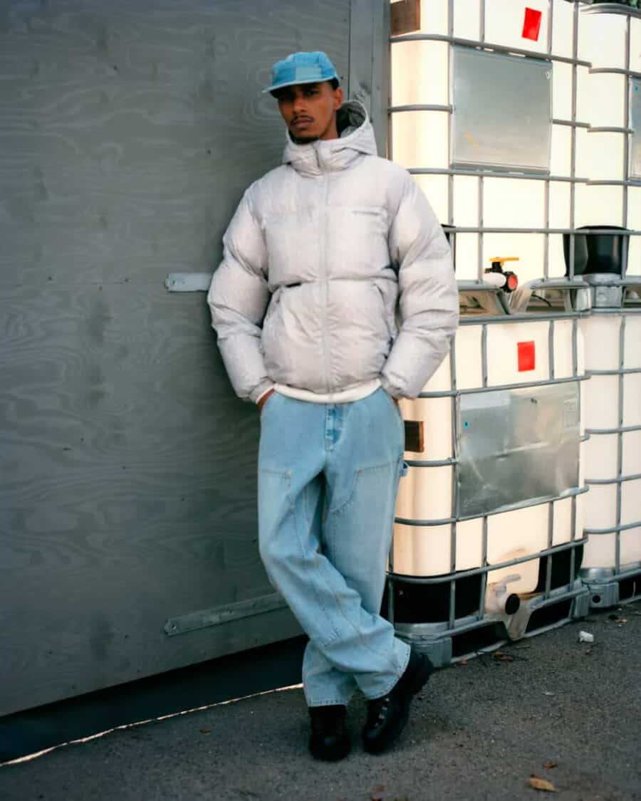 Man wearing Thisisneverthat streetwear baggy light wash jeans, light grey puffer jacket and light wash denim baseball cap with black boots