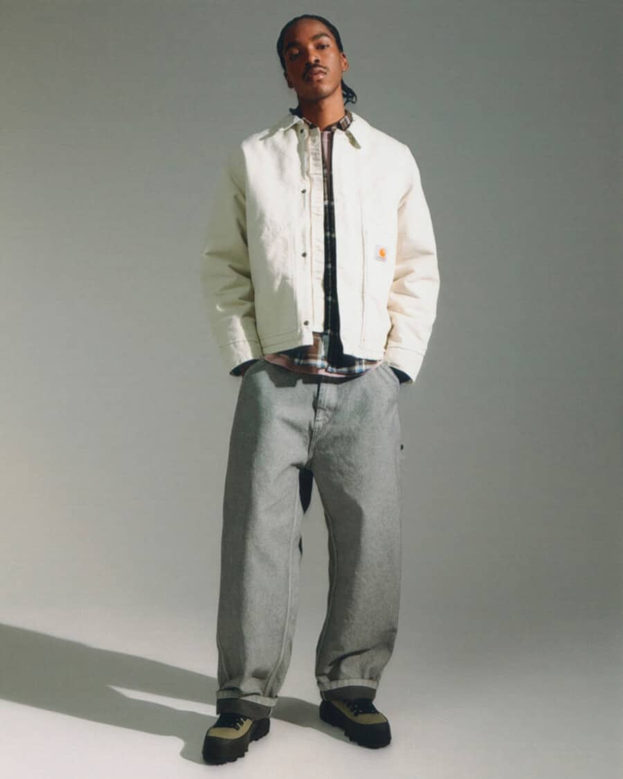Man wearing Carhartt WIP streetwear baggy grey jeans, brown check shirt, white worker jacket and green-black rubber boots
