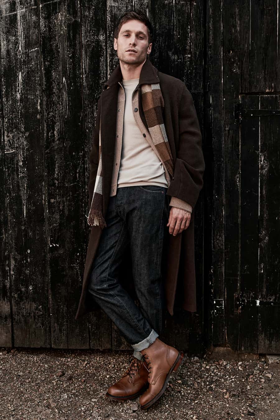 Men's raw denim jeans worn with brown boots, lightweight sweater and brown overcoat outfit