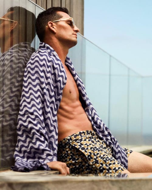Man sitting poolside wearing luxury printed Frescobol Carioca swim shorts and patterned towel over shoulders