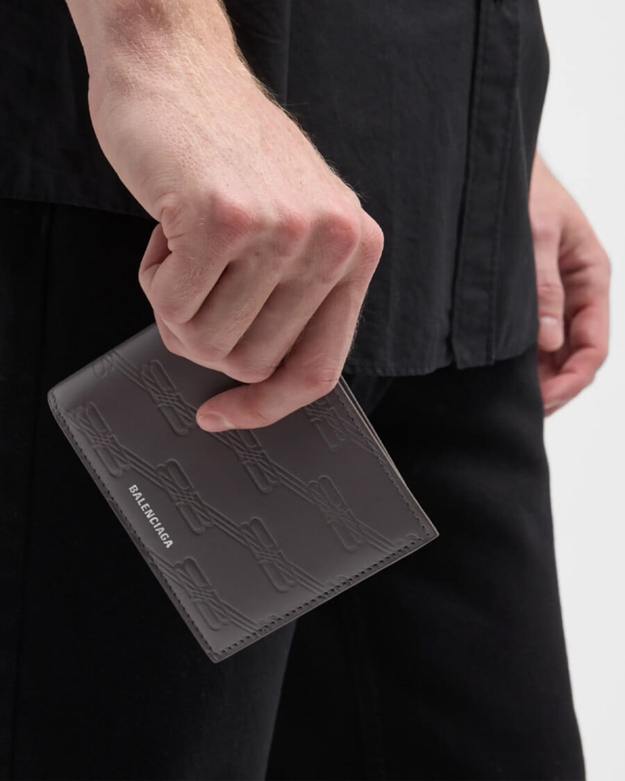 Man in black shirt and black pants holding a monogrammed Balenciaga logo wallet in brown leather