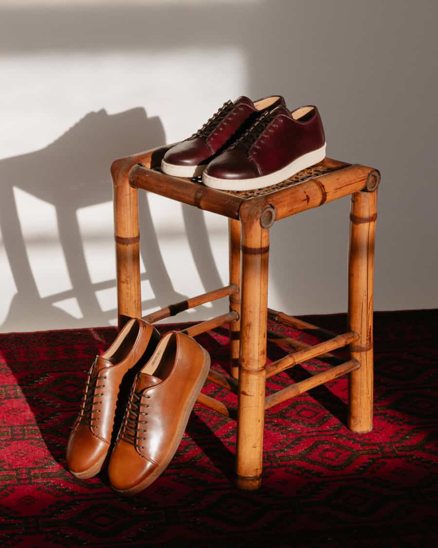Two pairs of handmade luxury men's sneakers set against a wooden stool