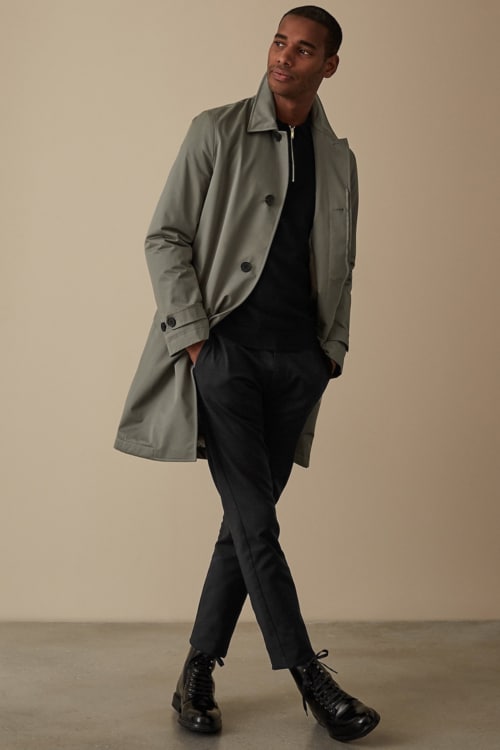 Men's black pleated trousers, black zip neck jumper, green trench coat and black boots outfit