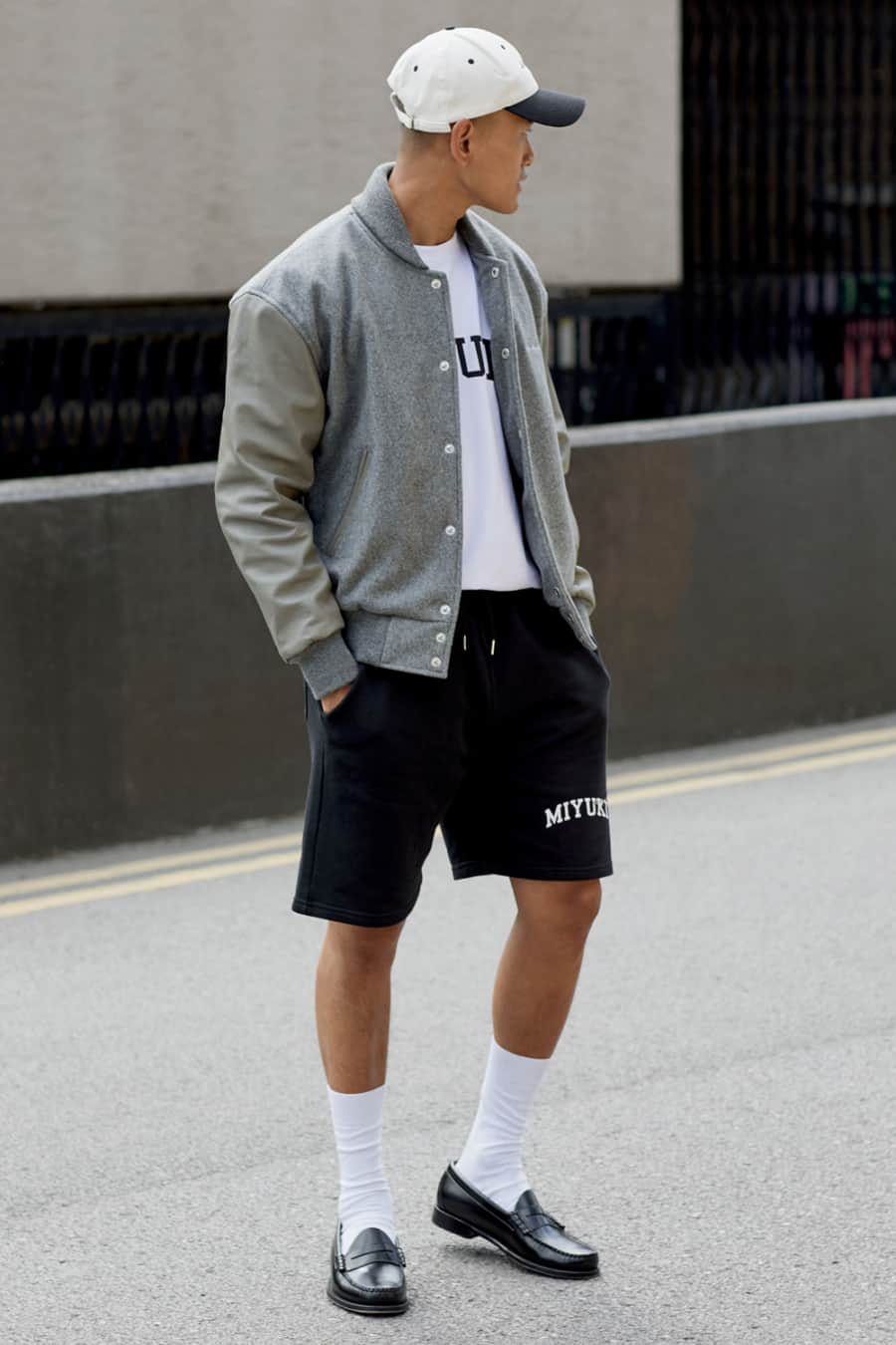Men's black shorts, white T-shirt, grey varsity jacket, white socks and black penny loafers outfit