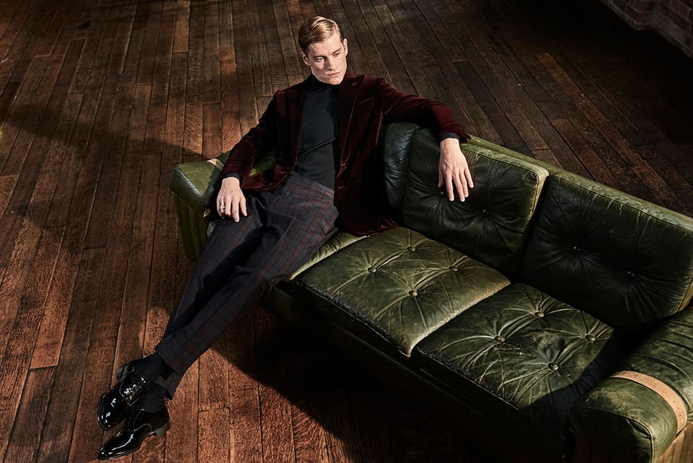 Man sat on sofa wearing semi formal dress: trousers, roll neck and velvet blazer