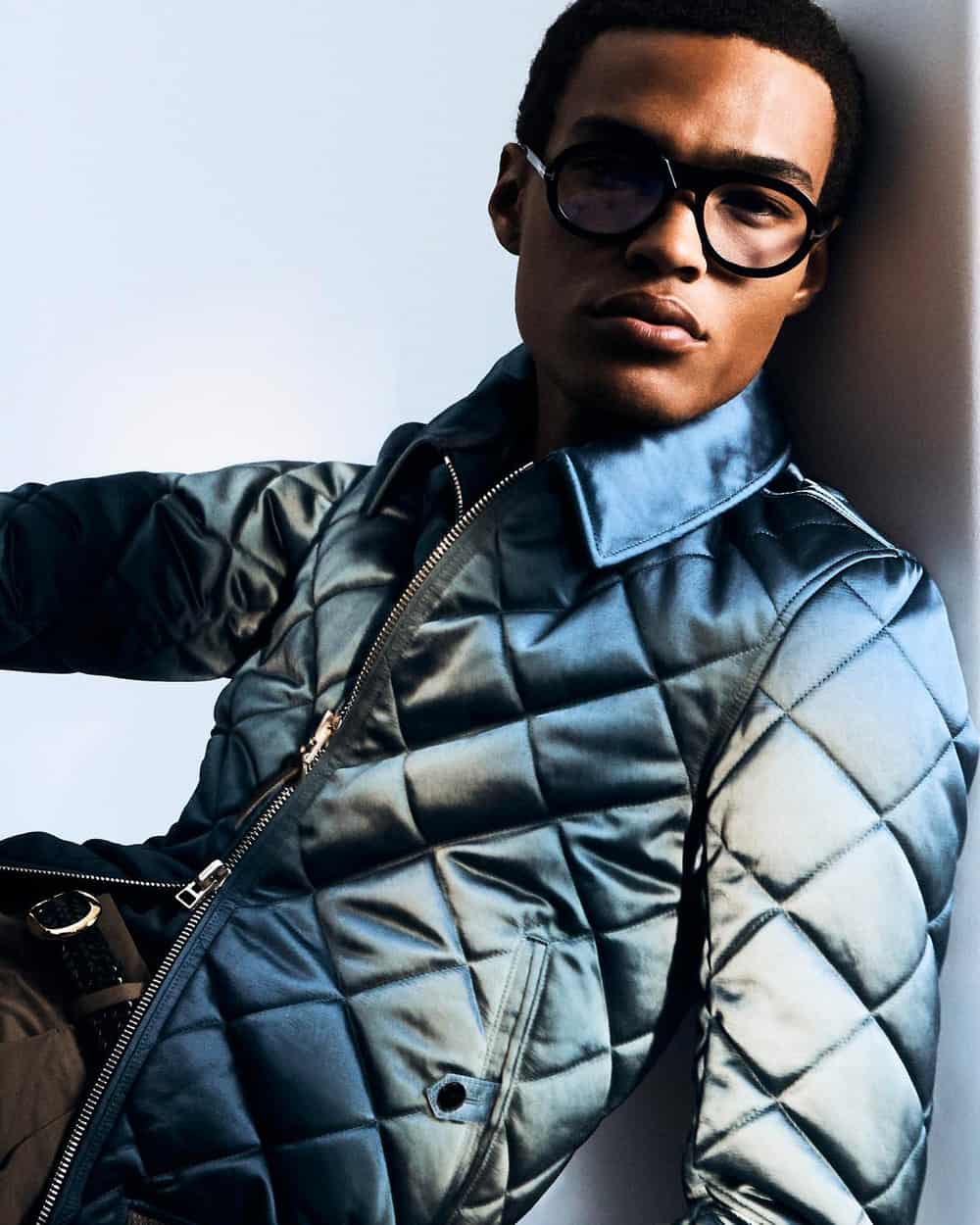 Black man wearing grey pants, light blue quilted jacket and black round lens spectacles by Tom Ford