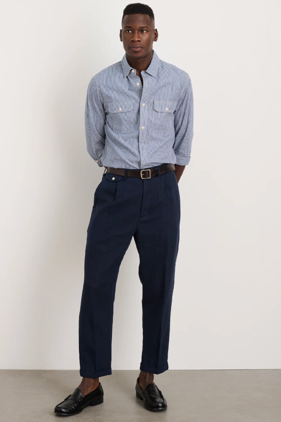 Men's cropped pleated navy pants, blue/white stripe workwear shirt, brown leather belt and sockless black leather penny loafers business casual outfit
