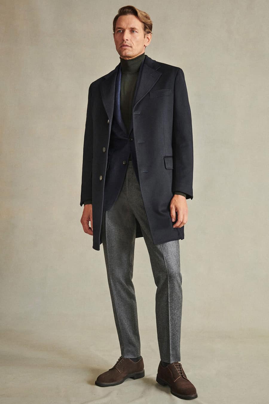 Men's grey wool trousers, green roll neck, navy blazer, navy overcoat and brown suede shoes