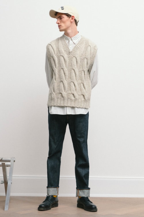 Men's raw denim jeans, white shirt and white cable knit sweater vest with baseball cap outfit