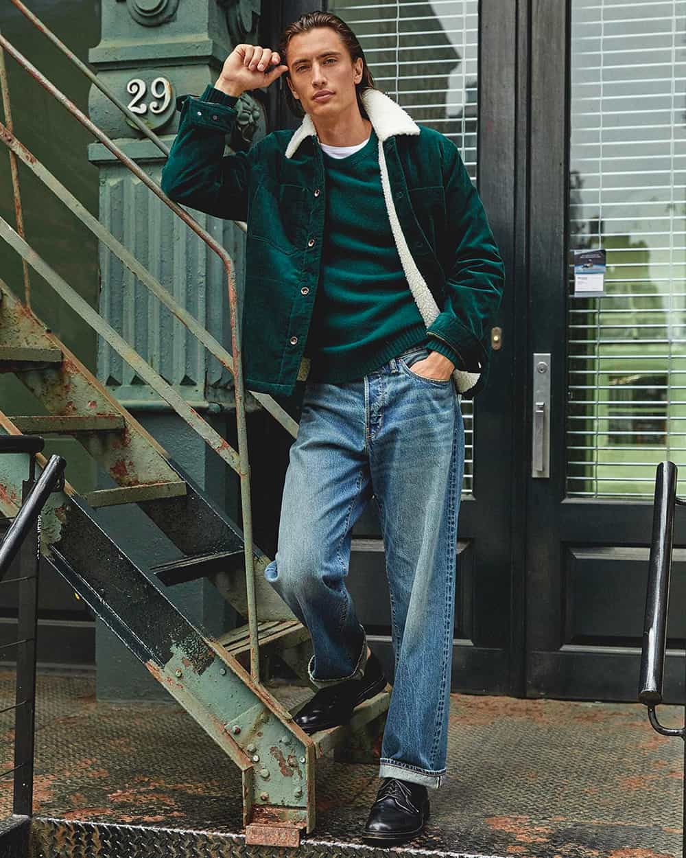 Man wearing Todd Snyder light wash jeans, green sweater, green borg collar jacket and black boots