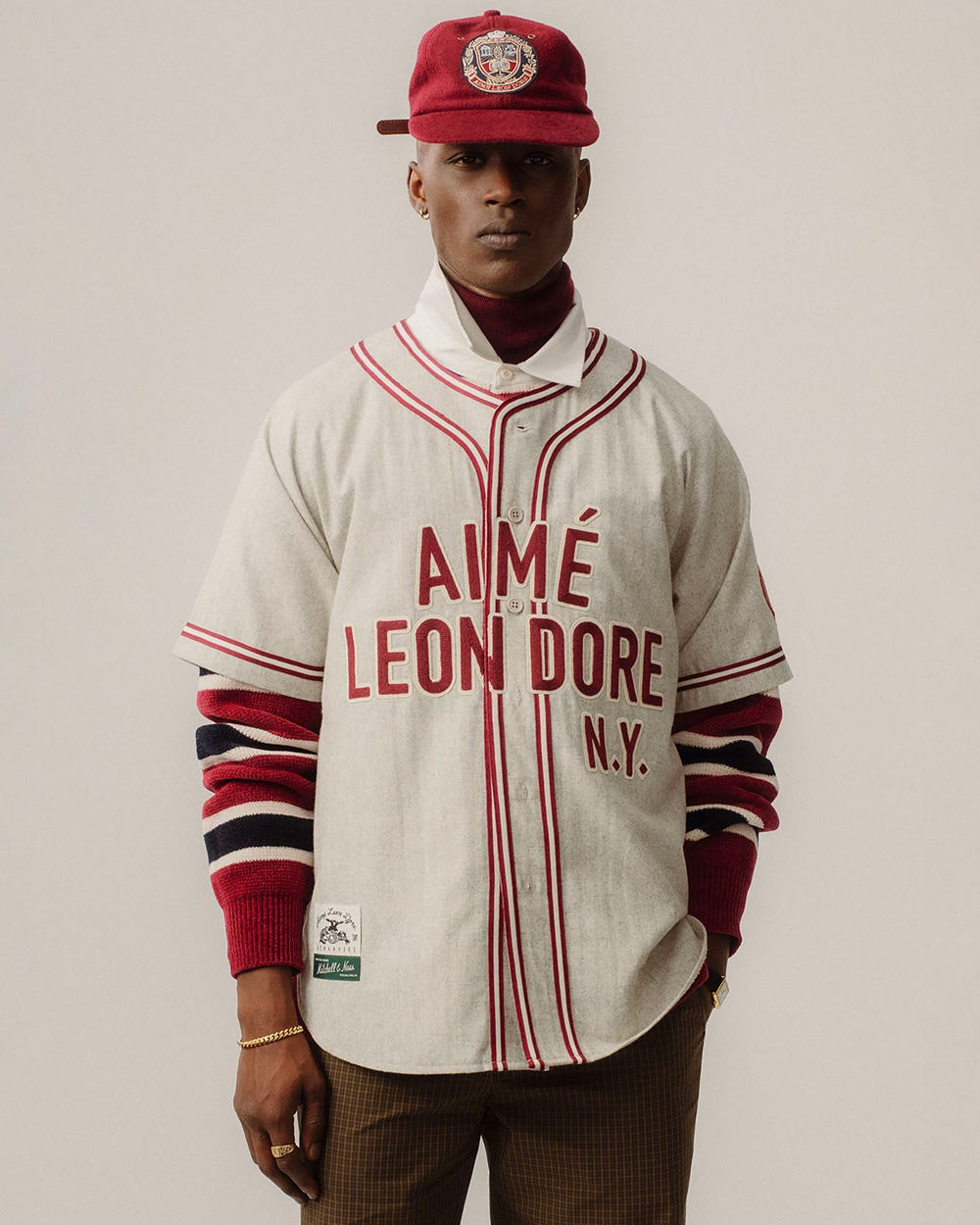 Black man wearing Aime Leon Dore brown pants, white shirt, white/red baseball top and red baseball cap