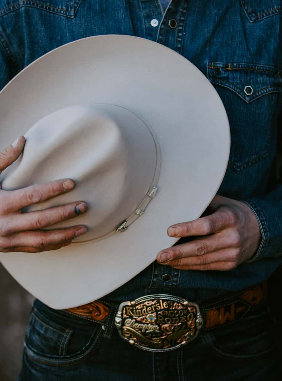 Cowboy wearing a denim shirt, jeans and big belt buckle holding a white felt cowboy hat