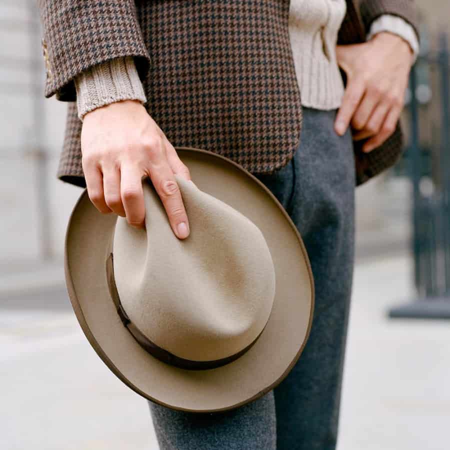 Man wearing grey wool trousers, knitted sweater and brown wool check blazer holding a brown felt fedora hat