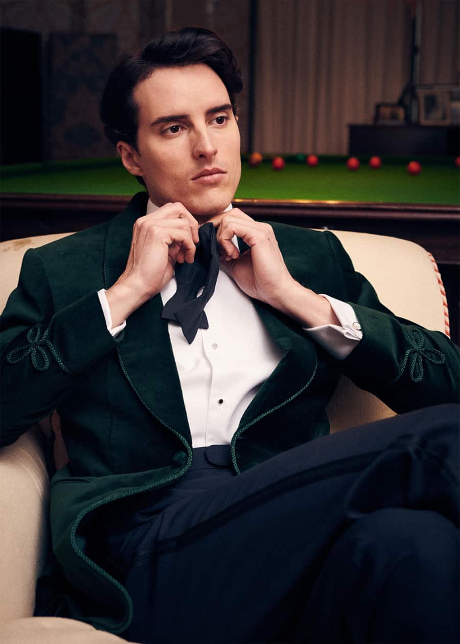 Man doing up his bow tie while wearing a white shirt and green velvet smoking jacket