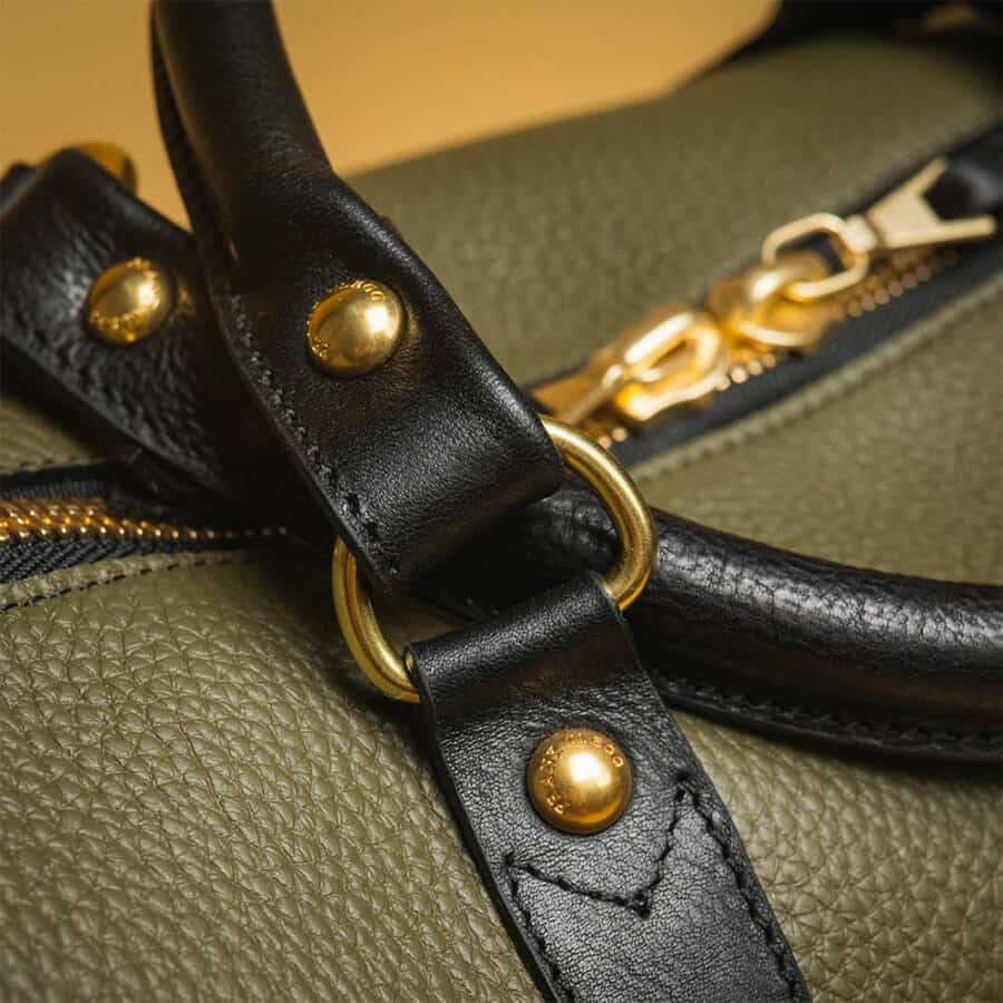 A close up of the brass metal hardware and black leather handles of a full grain green leather weekender bag