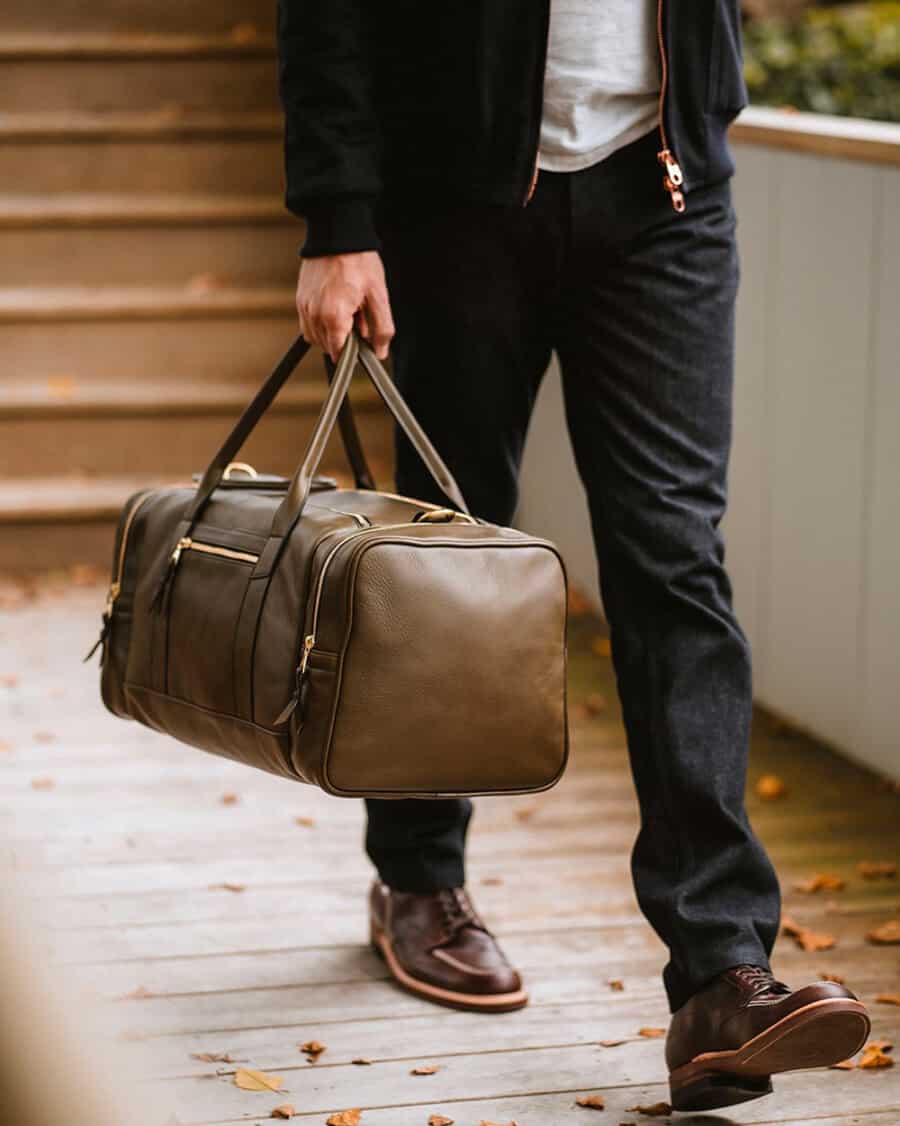 Man wearing raw denim jeans, grey T-shirt and black bomber jacket carrying a luxury green leather holdall bag