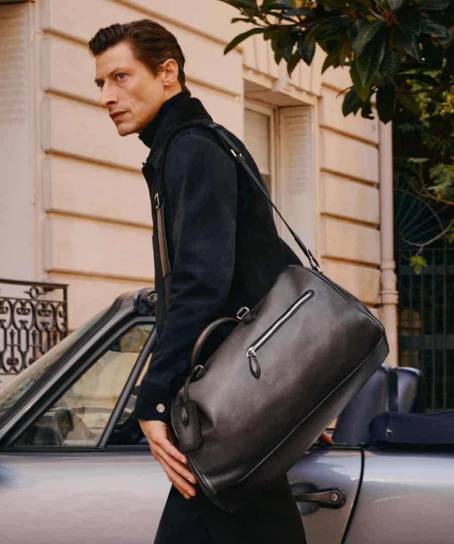 Man carrying luxury leather weekender bag over his shoulder wearing a suede jacket and turtleneck
