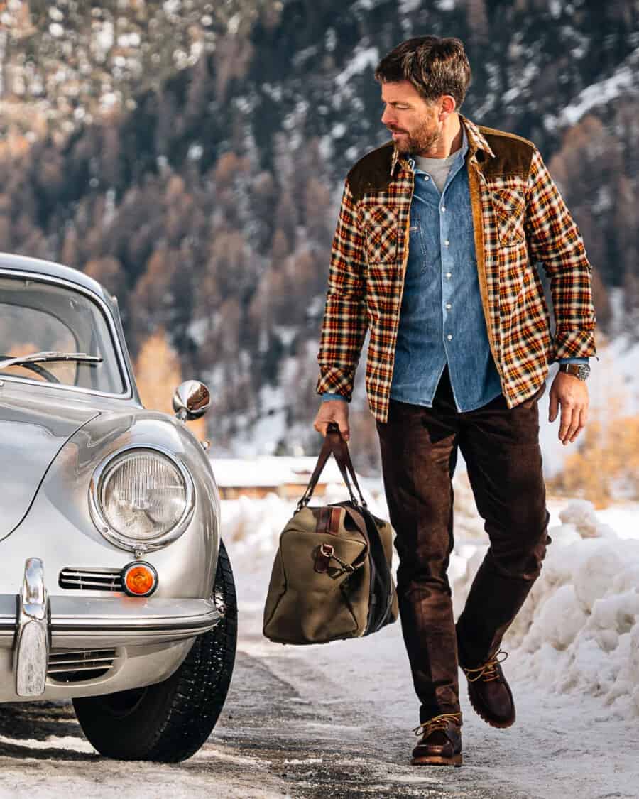 Man wearing brown chinos, white T-shirt, denim shirt, flannel overshirt and winter boots outside in the snow
