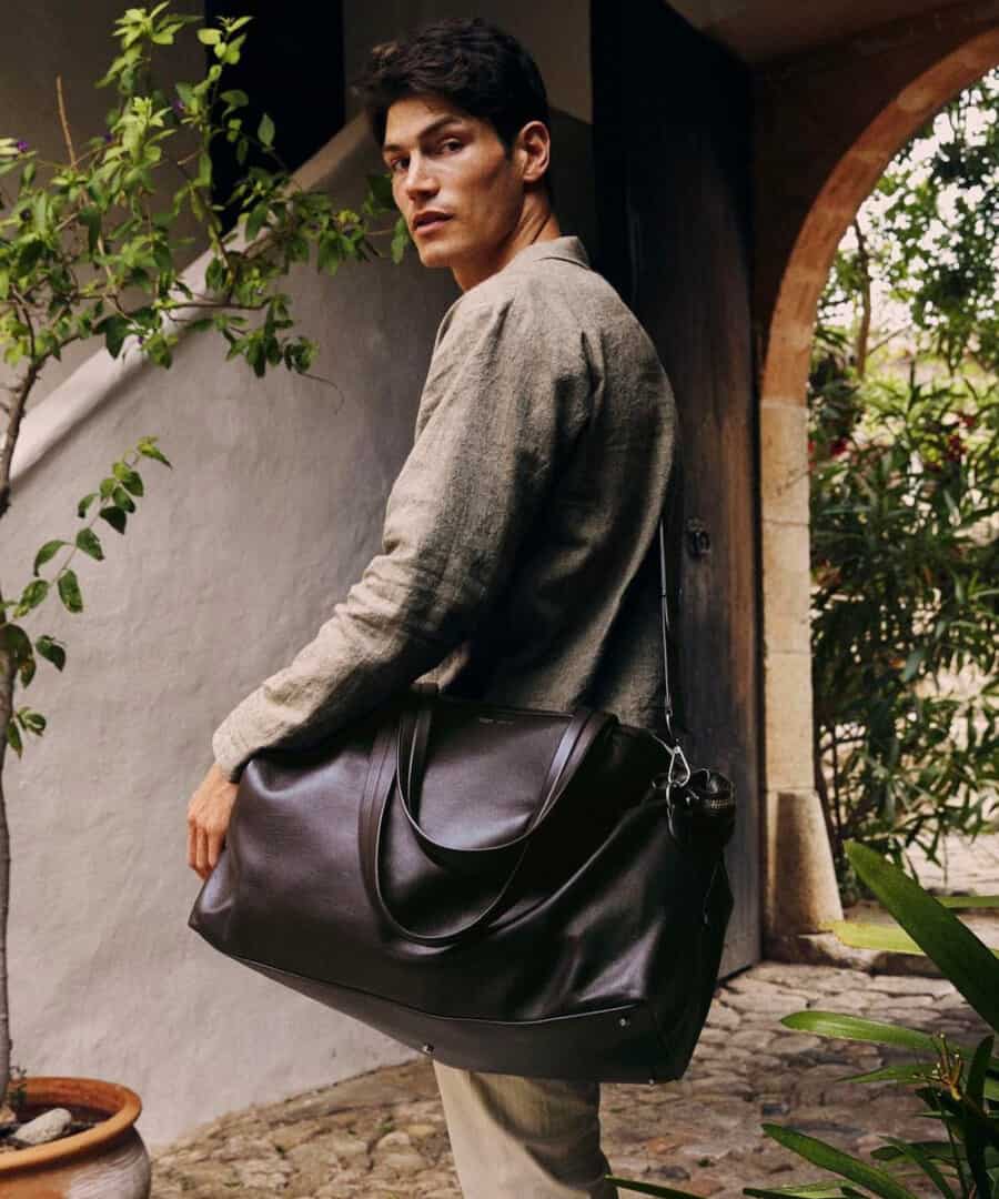 Man in linen shirt and trousers carrying a luxury leather weekender bag using the shoulder strap