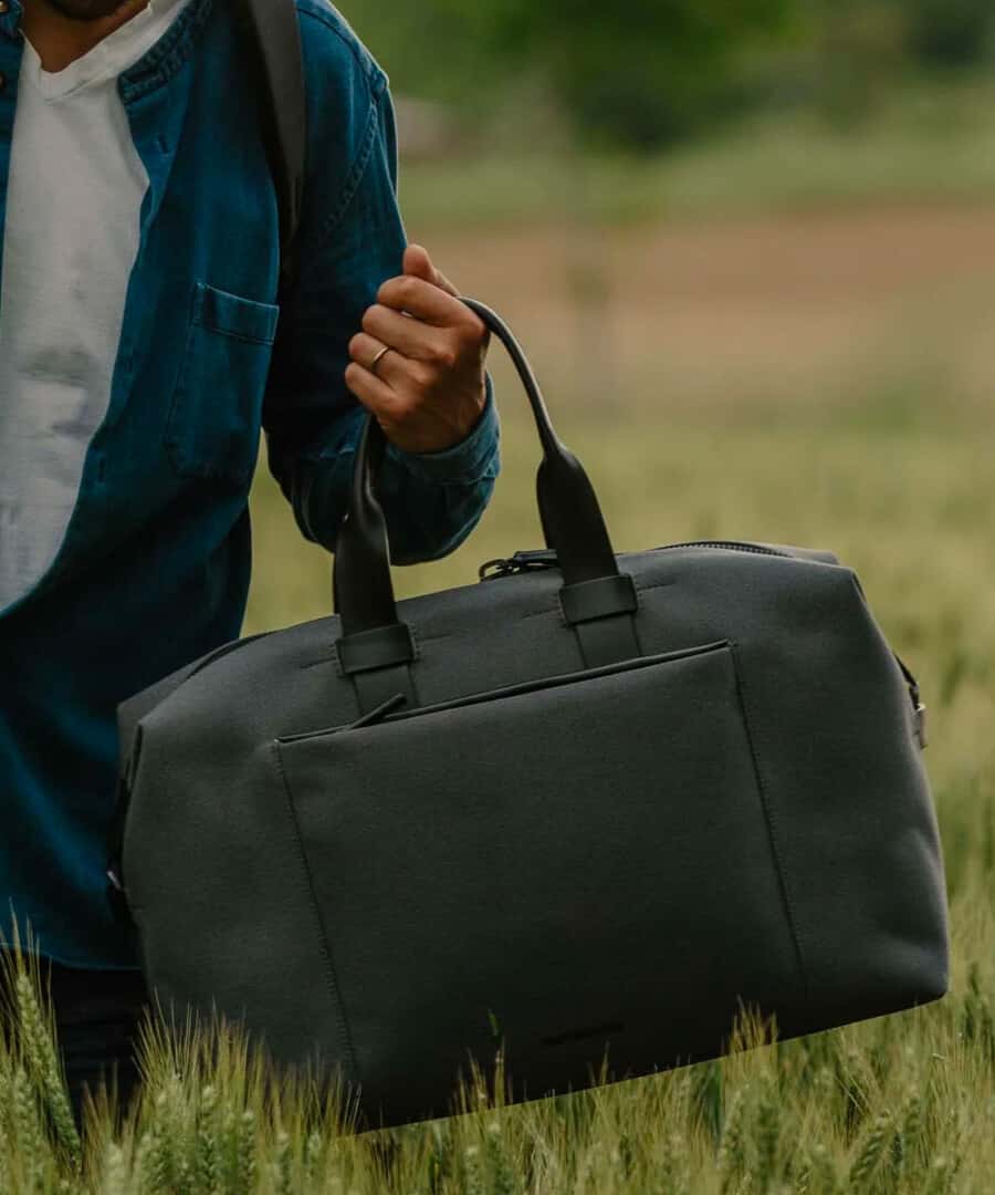Man outside in field holding black weekender bag by the handles