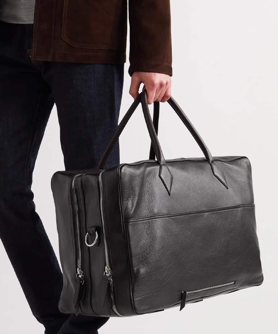 Man wearing navy trousers and brown suede jacket holding a black leather weekender bag by the handles