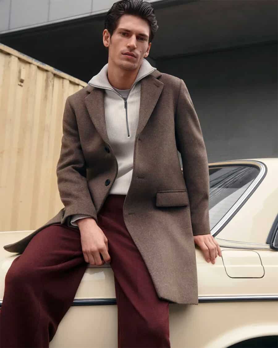 Man wearing burgundy trousers, ecru zip neck sweater and brown overcoat outfit
