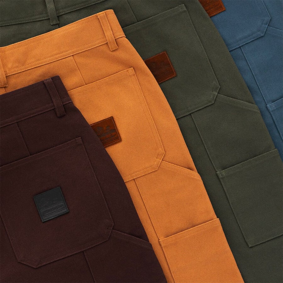 Four pairs of men's carpenter pants in brown, tan, green and blue laid out on top of each other