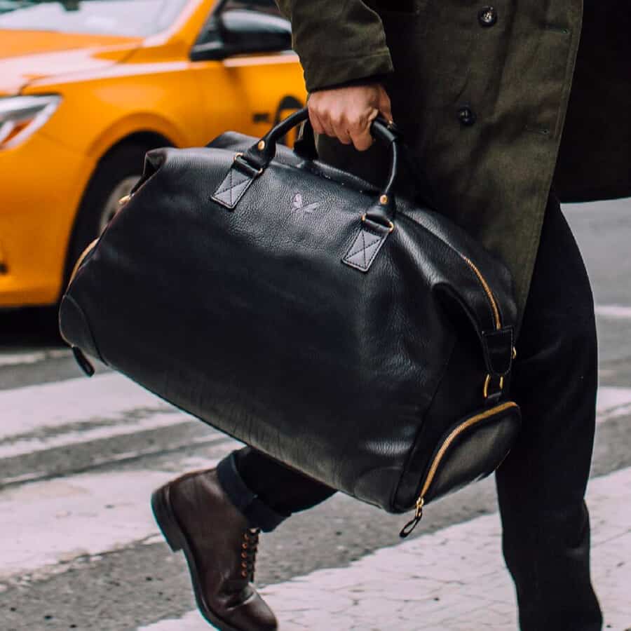 Man wearing black jeans and a green rain coat carrying a luxury black grained leather weekender bag