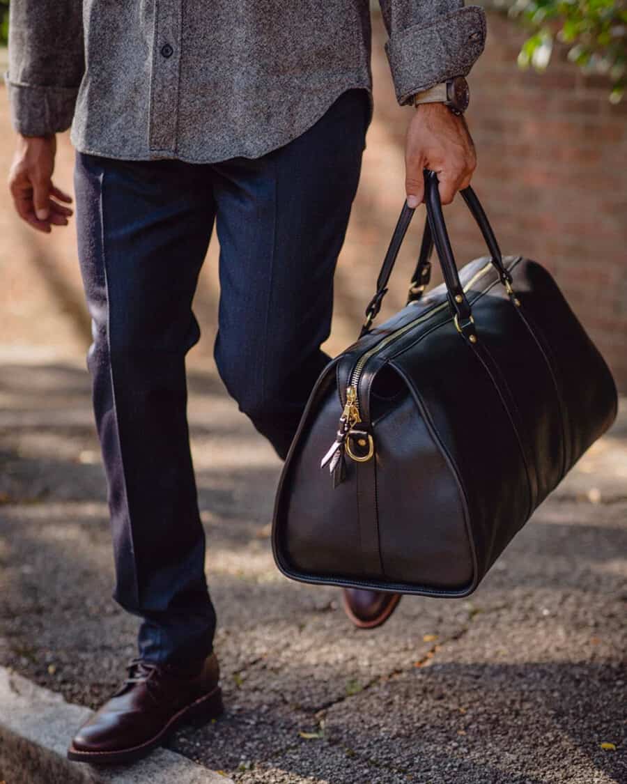 Man wearing navy trousers, grey flannel shirt and brown leather boots carrying a luxury brown leather weekender bag