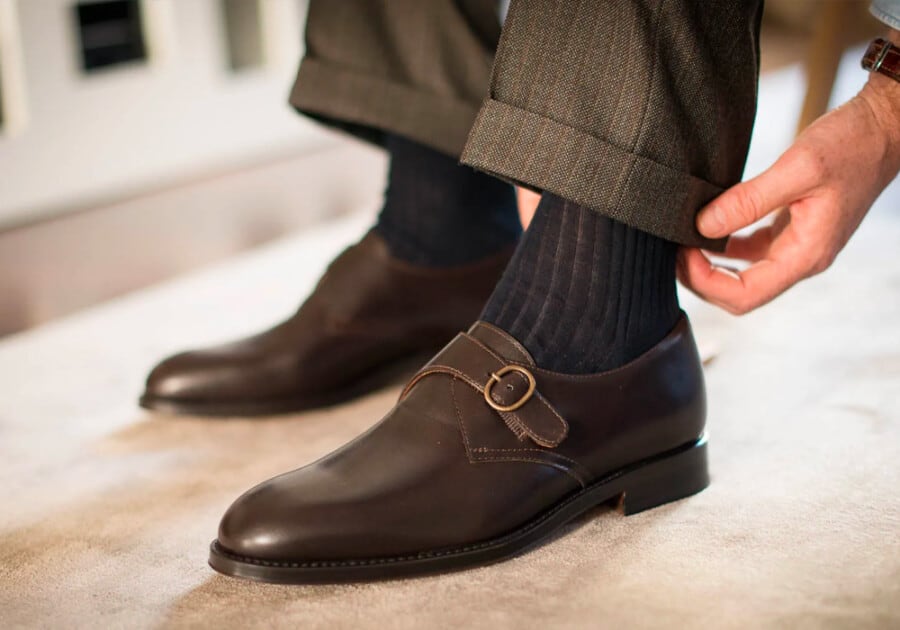 A pair of men's business casual monk strap shoes in brown leather worn with navy socks and grey turn up trousers
