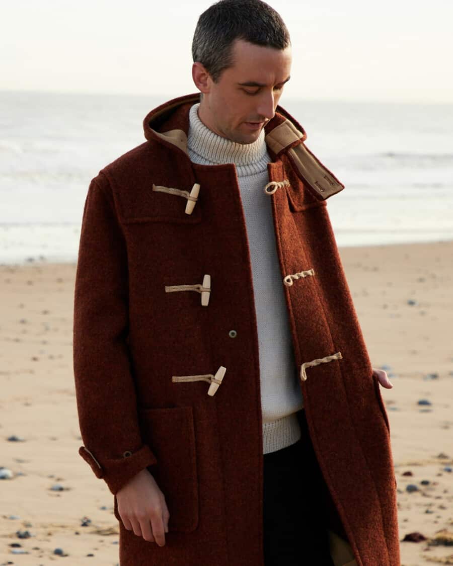 Man wearing a brown duffle coat over a white turtleneck jumper on a cold beach