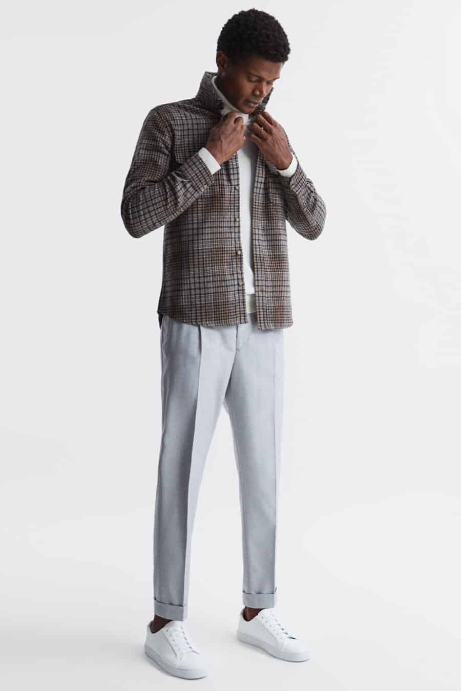 Men's light grey pleated trousers, white roll neck, brown checked flannel overshirt and white sneakers outfit