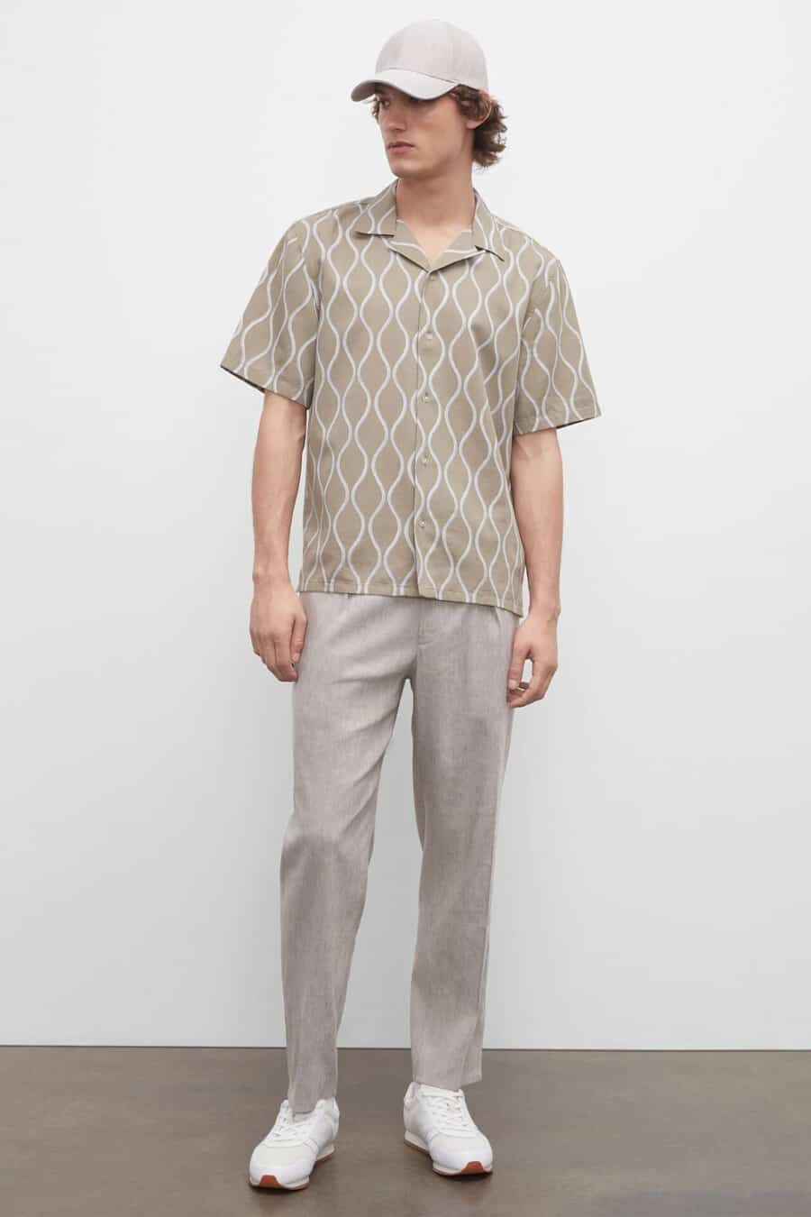 Men's light grey linen trousers, patterned brown short sleeve shirt, white sneakers and light grey baseball cap outfit