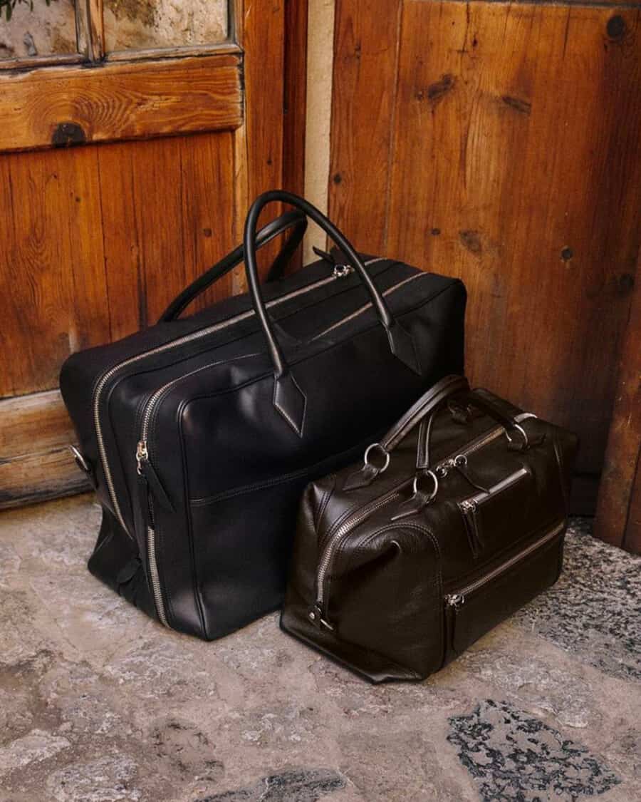 Two luxury black leather holdall bags set on floor against wooden doors