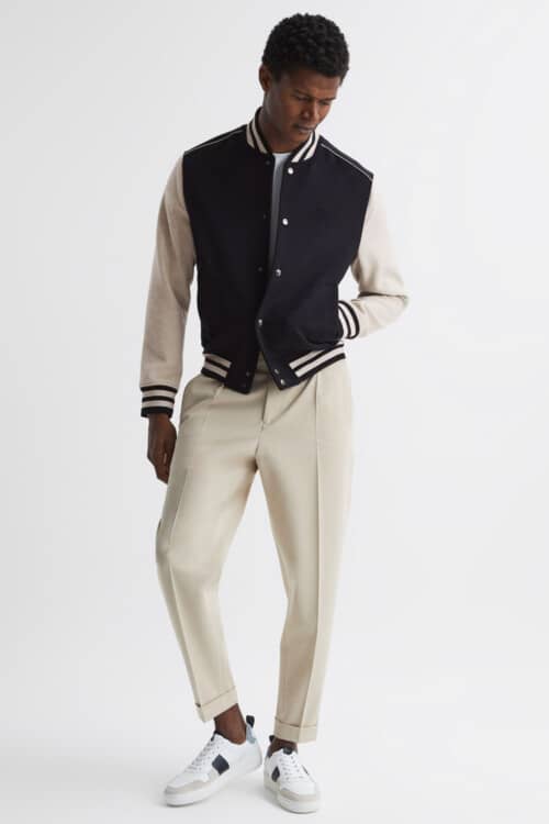 Men's tailored cream trousers, navy varsity jacket, white T-shirt and white sneakers outfit
