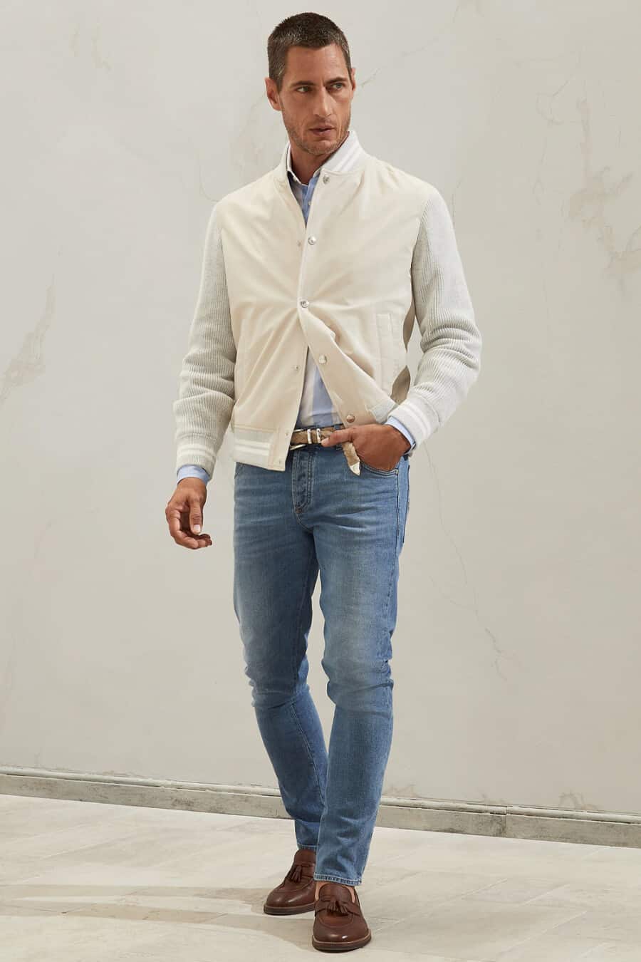Men's mid-wash jeans, striped shirt, cream varsity jacket and brown leather tassel loafers outfit