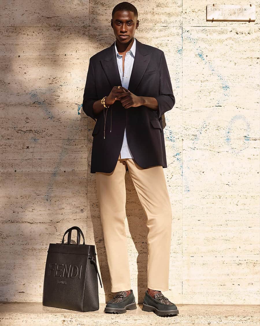 Man wearing khaki pants, blue shirt, navy blazer, chunky loafers with a luxury leather Fendi tote bag sitting on floor next to him
